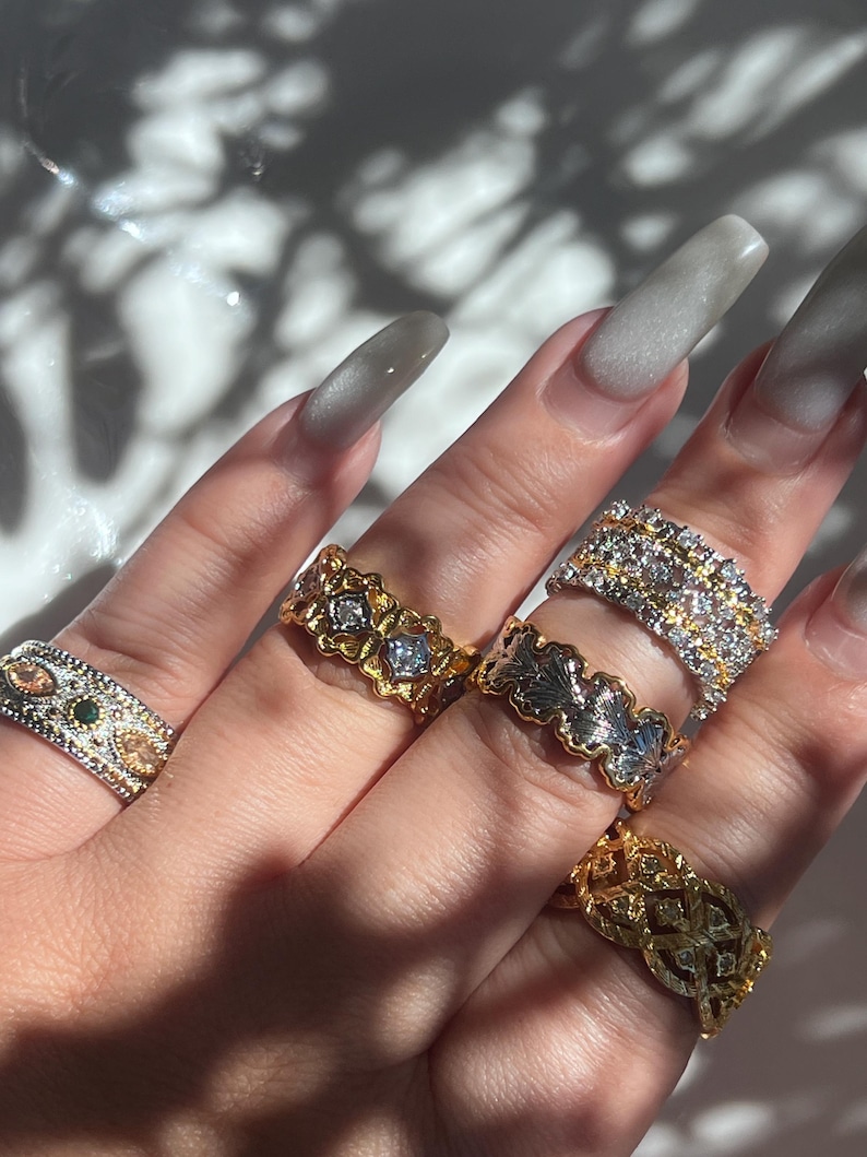 May include: A hand displaying several ornate rings. The rings feature various designs, including floral patterns, geometric shapes, and gemstone accents. The rings are gold and silver-toned, with clear and colored stones. The nails are long and painted a light gray color.