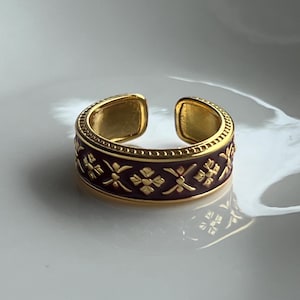 Puede incluir: Un anillo abierto de tono dorado con una incrustación de esmalte color burdeos intenso. La banda está decorada con un patrón repetitivo de motivos florales geométricos dorados en relieve y tiene un borde de cuentas en la parte superior e inferior.
