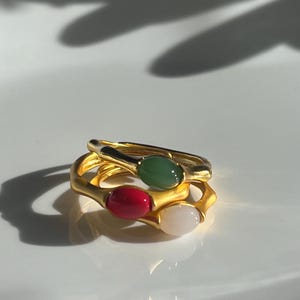 Pu&ograve; includere: Tre anelli dorati con pietre preziose ovali rosse, verdi e bianche. Gli anelli sono impilati, con le pietre preziose rivolte verso l'alto. Gli anelli hanno un design strutturato e organico.