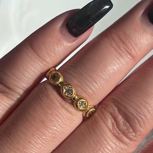 May include: A gold-colored ring with a repeating pattern of diamond-shaped settings, each containing a small, clear gemstone. The ring has a textured, rope-like design along the band. The ring is shown on a person's finger.
