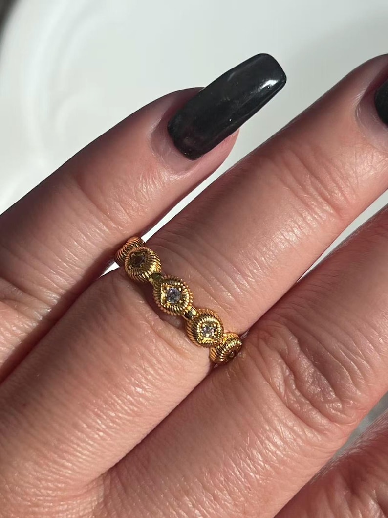 May include: A gold-colored ring with a repeating pattern of diamond-shaped settings, each containing a small, clear gemstone. The ring has a textured, rope-like design along the band. The ring is shown on a person's finger.