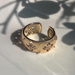 May include: A gold-coloured adjustable ring with a floral design. The ring features a textured band with vertical lines and small, round embellishments. The open design allows for size adjustment. The ring is on a white surface.