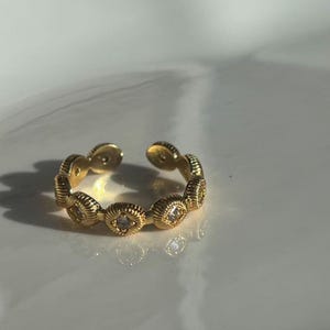 May include: Gold-toned adjustable ring featuring a series of oval-shaped, textured links. Each link is adorned with a small, clear gemstone. The ring's design is open, allowing for size adjustment. The ring is displayed on a white surface.