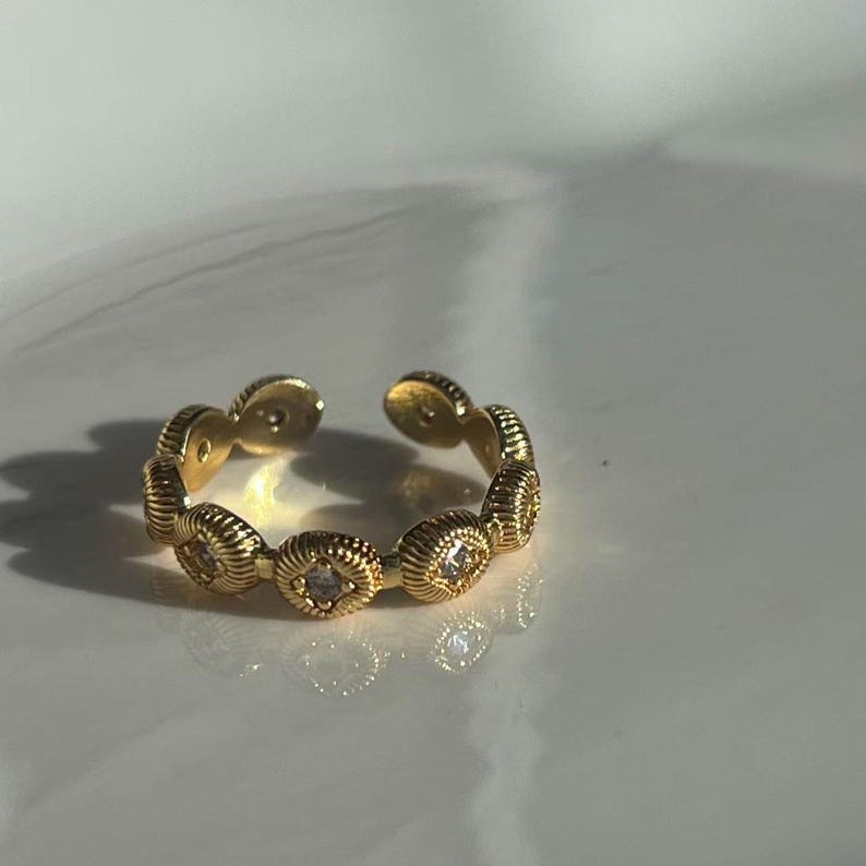 May include: Gold-toned adjustable ring featuring a series of oval-shaped, textured links. Each link is adorned with a small, clear gemstone. The ring's design is open, allowing for size adjustment. The ring is displayed on a white surface.