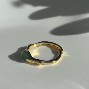 Pu&ograve; includere: Un anello dorato con una fascia in stile bamb&ugrave; e una pietra rotonda verde chiaro. L'anello &egrave; posto su uno sfondo bianco, con ombre che aggiungono profondit&agrave;. &Egrave; un gioiello.