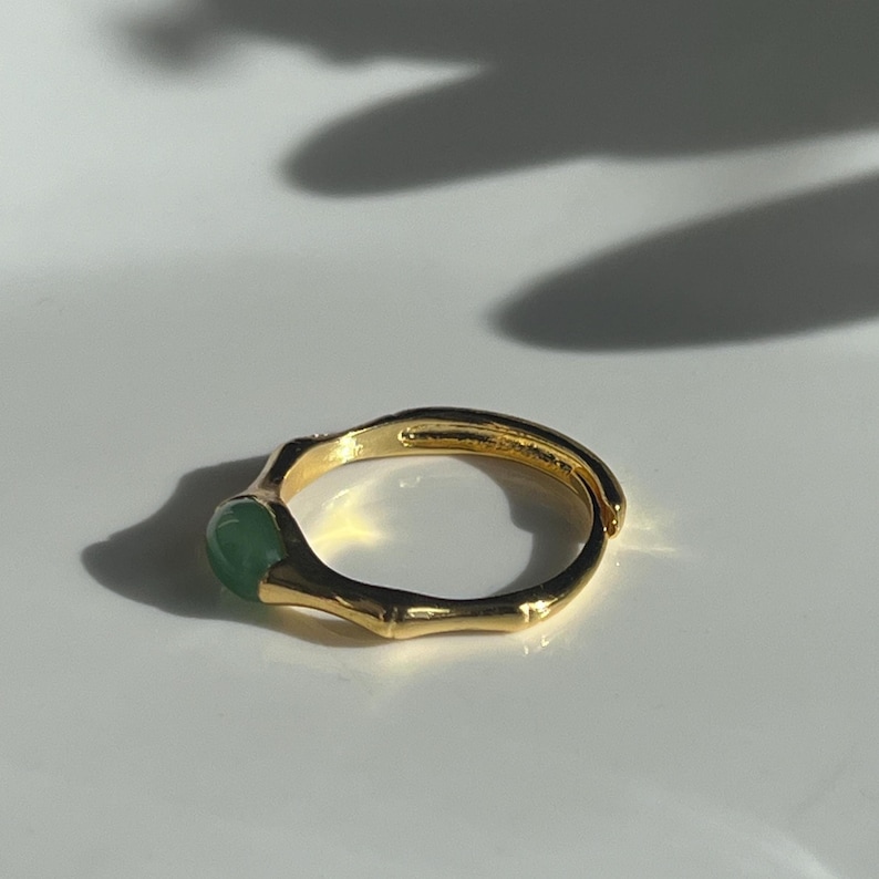 Pu&ograve; includere: Un anello dorato con una fascia in stile bamb&ugrave; e una pietra rotonda verde chiaro. L'anello &egrave; posto su uno sfondo bianco, con ombre che aggiungono profondit&agrave;. &Egrave; un gioiello.