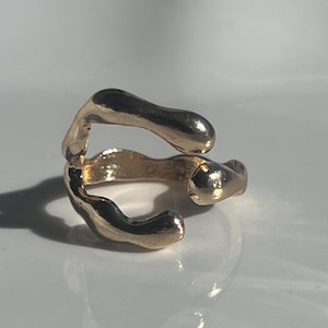 Peut inclure: Une bague dorée à la forme ouverte, irrégulière et organique. L'anneau présente une surface lisse et réfléchissante avec des extrémités arrondies et bulbeuses qui ne se rejoignent pas, créant un aspect fluide et métallique brillant.
