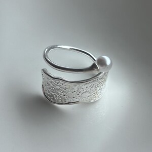 May include: A silver-toned open ring featuring a textured, hammered-effect band that wraps around the finger. One end of the band terminates in a smooth, round white pearl, while the other end forms a wider, irregular, and bumpy surface.