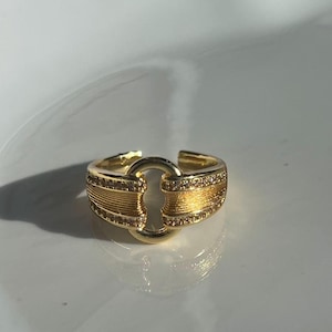 May include: A gold ring with a distinctive design. The ring has a central circular feature linked to two rectangular bands. The bands are decorated with small, sparkling stones and horizontal lines.