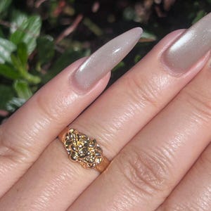 May include: A gold-colored ring with a textured, nugget-like design. The ring is worn on a finger with long, polished nails. The ring's design features a cluster of irregular shapes, creating a unique and eye-catching piece of jewelry.