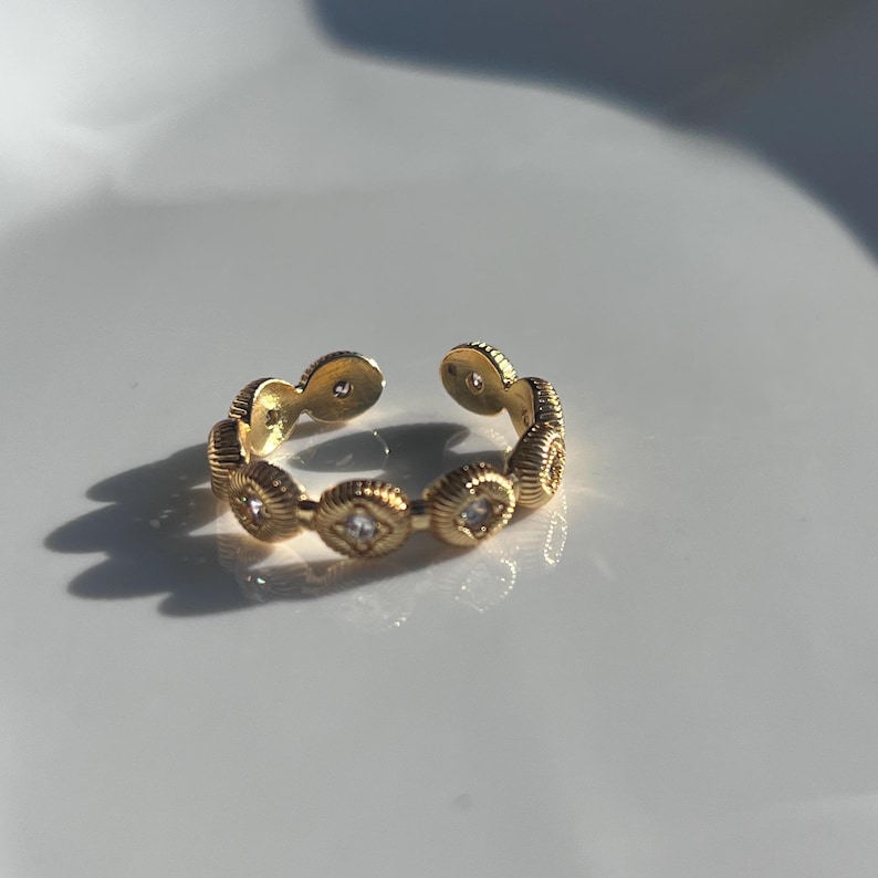 May include: A gold-colored adjustable ring with a repeating pattern of textured, circular elements. Each circular element is adorned with a small, square-shaped, clear gemstone. The ring is open at the top.