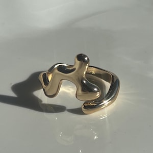 May include: A gold-colored ring with an abstract, organic design. The ring features a flowing, asymmetrical shape with rounded edges and a smooth, polished finish. The ring is set against a white background.