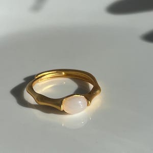 Pu&ograve; includere: Un anello dorato con una pietra ovale rosa chiaro. L'anello ha un design unico e organico con una fascia strutturata. La pietra &egrave; incastonata in una lunetta. L'anello &egrave; esposto su una superficie bianca.