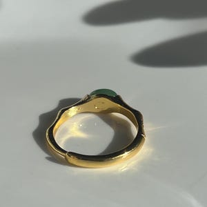Pu&ograve; includere: Un anello color oro con una fascia strutturata e una piccola gemma verde rotonda. L'anello &egrave; posto su uno sfondo bianco, con ombre che aggiungono profondit&agrave;. Il design dell'anello &egrave; organico ed elegante.