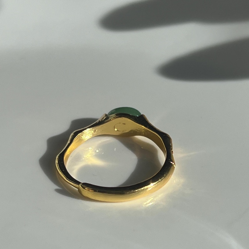 Pu&ograve; includere: Un anello color oro con una fascia strutturata e una piccola gemma verde rotonda. L'anello &egrave; posto su uno sfondo bianco, con ombre che aggiungono profondit&agrave;. Il design dell'anello &egrave; organico ed elegante.
