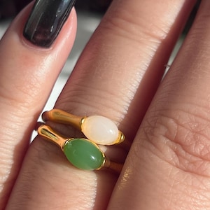 Pu&ograve; includere: Due anelli dorati, ciascuno con una gemma ovale. Un anello ha una pietra rosa pallido, l'altro una pietra verde chiaro. Gli anelli sono mostrati su un dito, evidenziando il design e il colore dei gioielli.