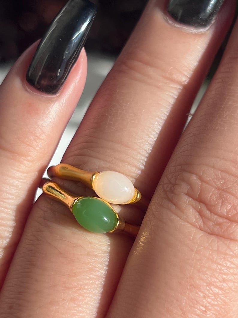 Pu&ograve; includere: Due anelli dorati, ciascuno con una gemma ovale. Un anello ha una pietra rosa pallido, l'altro una pietra verde chiaro. Gli anelli sono mostrati su un dito, evidenziando il design e il colore dei gioielli.