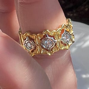 May include: A gold ring with a repeating floral design. Each floral element features a central clear stone. The ring is held by a person's finger, with the sunlight reflecting off the ring's surface.