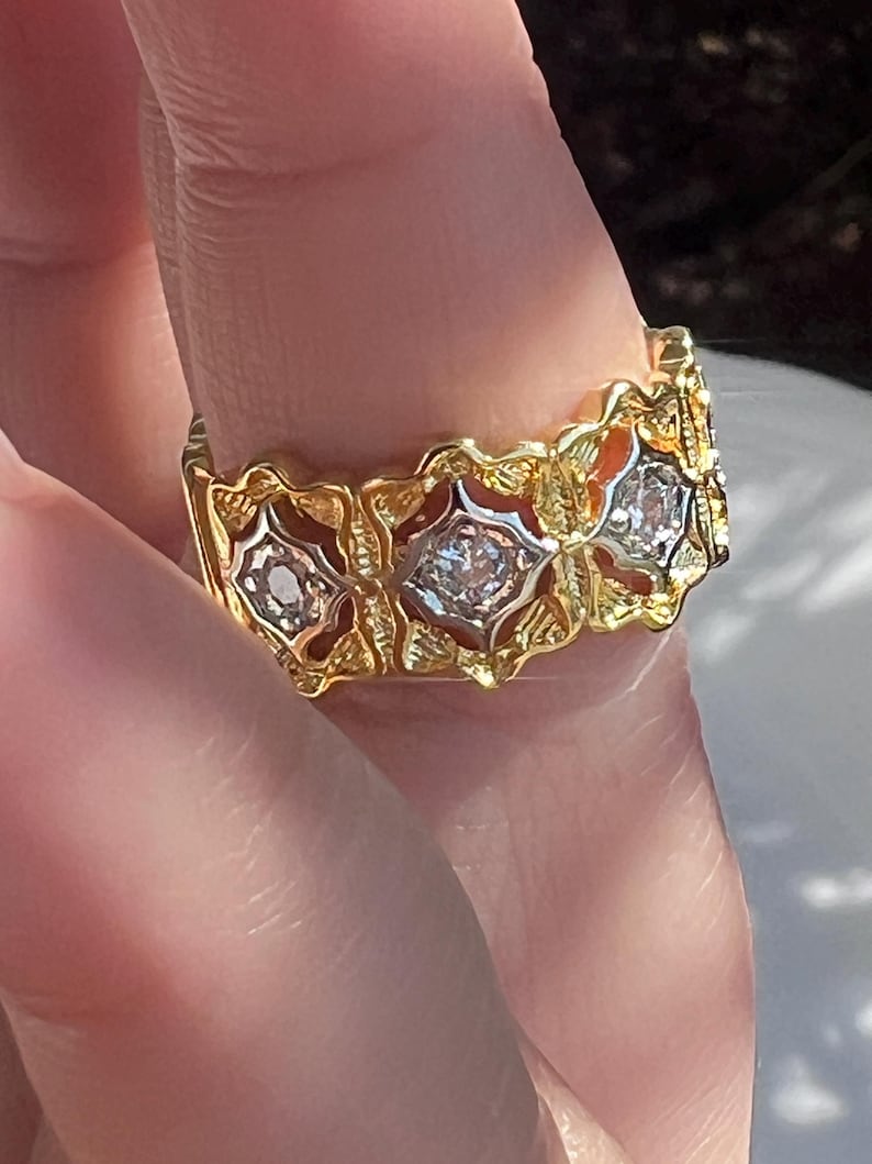 May include: A gold ring with a repeating floral design. Each floral element features a central clear stone. The ring is held by a person's finger, with the sunlight reflecting off the ring's surface.