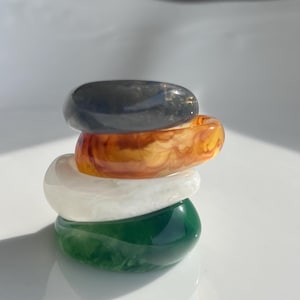 May include: Four resin rings stacked on top of each other. The rings are in shades of dark grey, orange, white, and green. The rings are made of resin and are stacked on a white surface. The rings are approximately 2 cm wide.
