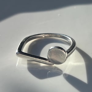 Könnte beinhalten: Ein silberner Ring mit modernem Design. Der Ring hat eine kreisförmige Scheibe und einen rechteckigen Stab, die mit einem gebogenen Band verbunden sind. Der Ring besteht aus einem glänzenden, metallischen Material.