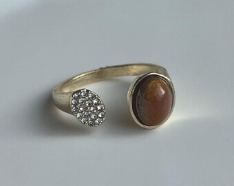 May include: A gold-toned open ring featuring a brown oval gemstone and a cluster of small, clear crystals. The ring has a split shank design, with the gemstone and crystal cluster on opposite ends.