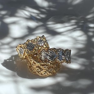 May include: Close-up of several gold rings with intricate floral and geometric designs. Some rings feature silver accents and small gemstones. The rings are stacked on top of each other, creating a layered effect. The background is a soft white surface.