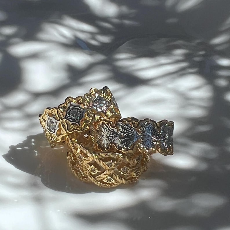 May include: Close-up of several gold rings with intricate floral and geometric designs. Some rings feature silver accents and small gemstones. The rings are stacked on top of each other, creating a layered effect. The background is a soft white surface.