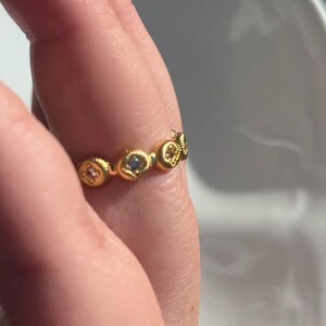 May include: A gold-colored ring with a repeating pattern of small, round, decorative elements. Each element features a small, colorful gemstone. The ring is worn on a finger.