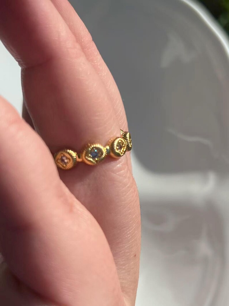 May include: A gold-colored ring with a repeating pattern of small, round, decorative elements. Each element features a small, colorful gemstone. The ring is worn on a finger.