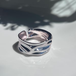 May include: A silver-toned open ring featuring a wide, irregular band with a polished, reflective surface and deep, flowing indentations that create a liquid-like appearance. The ring has a visible gap at the back, allowing for size adjustment.