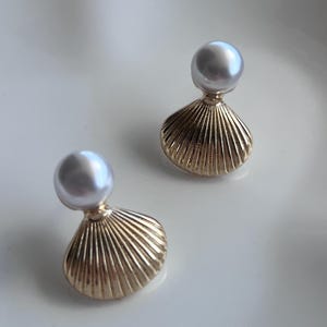 Op de afbeelding: Een paar oorknopjes met een gladde, ronde witte parel bevestigd aan een goudkleurige metalen basis in de vorm van een geribbelde schelp. Het ontwerp toont een gepolijste metalen afwerking op het schelpvormige deel en een glanzend pareloppervlak.