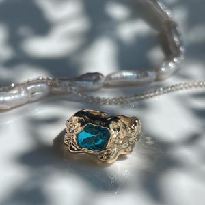 May include: A gold ring with a textured design, featuring a rectangular, blue gemstone. A strand of white pearls is in the background. The ring is displayed on a white surface, with shadows.