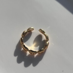 May include: A gold-colored open ring with a seashell design. The ring is made of a shiny metal and has a textured surface. The ring is open at the top and has a shadow on a white surface.