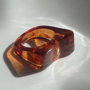 Peut inclure: Une bague épaisse fabriquée en résine translucide de couleur ambre, présentant une forme carrée arrondie et lisse avec des inclusions tourbillonnantes brun foncé dans tout le matériau qui captent la lumière et créent un effet chaleureux.