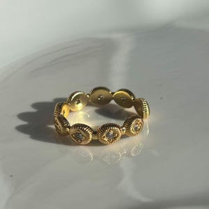May include: A gold-colored ring with a repeating pattern of textured, circular elements. Each circle features a small, clear gemstone at its center. The ring is set against a white background, with soft lighting.