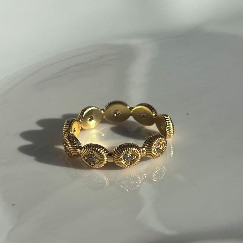 May include: A gold-colored ring with a repeating pattern of textured, circular elements. Each circle features a small, clear gemstone at its center. The ring is set against a white background, with soft lighting.