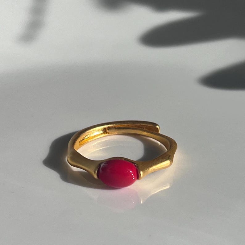 Pu&ograve; includere: Un anello color oro con una pietra rosa brillante a forma ovale. L'anello ha un design aperto unico con una finitura liscia e lucida. L'anello &egrave; posto su uno sfondo bianco, con ombre.