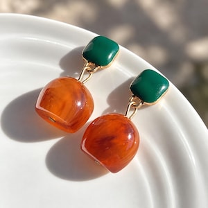 Peut inclure: Une paire de boucles d'oreilles pendantes composée d'un clou carré en émail vert foncé relié par un anneau en métal doré à une perle arrondie translucide de couleur ambre avec une texture marbrée. Finition polie et design pendant.