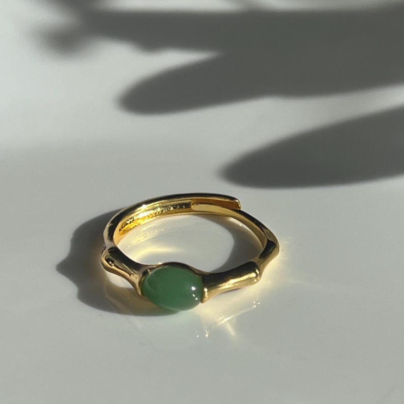 Pu&ograve; includere: Un anello color oro con una pietra verde a forma ovale. L'anello ha una fascia strutturata e un design semplice ed elegante. L'anello &egrave; esposto su una superficie bianca.