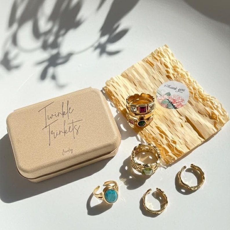 Pu&ograve; includere: Una collezione di anelli dorati con vari design e pietre preziose, tra cui turchese, smeraldo e rubino. Gli anelli sono esposti con una piccola scatola beige con la scritta "Twinkle Trinkets" e carta decorativa. &Egrave; presente anche un adesivo con arte floreale.