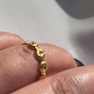 May include: A gold ring with a repeating pattern of small, textured, oval-shaped designs. Each oval has a small, clear stone in the center. The ring is worn on a finger.