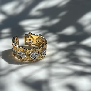 May include: A gold-toned adjustable ring with a floral and geometric design. The ring features alternating sections of gold and silver-toned metal, with small, clear gemstones set within the silver sections. The ring is open at the top.