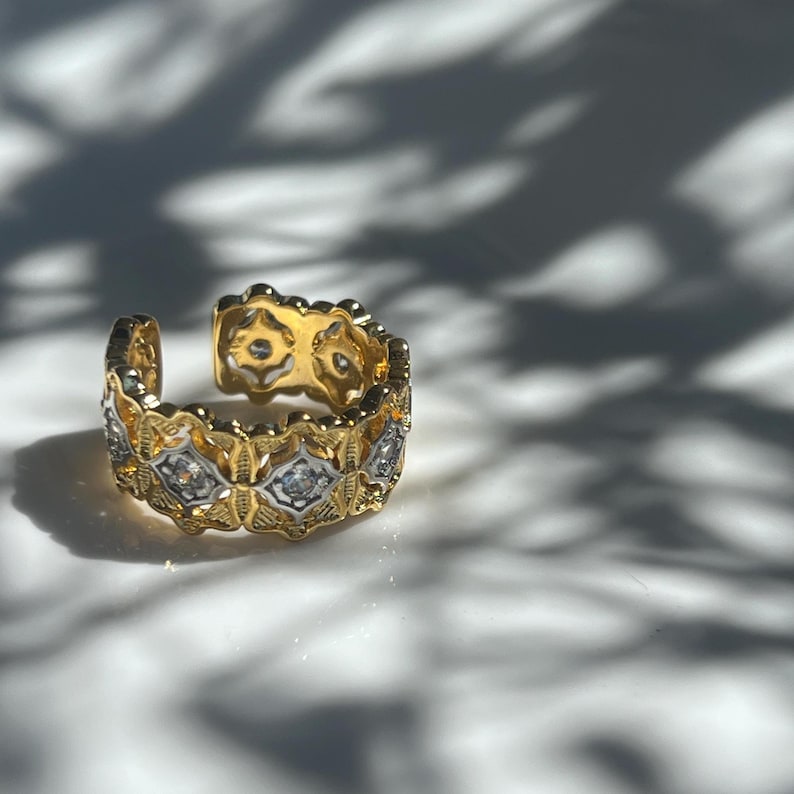 May include: A gold-toned adjustable ring with a floral and geometric design. The ring features alternating sections of gold and silver-toned metal, with small, clear gemstones set within the silver sections. The ring is open at the top.