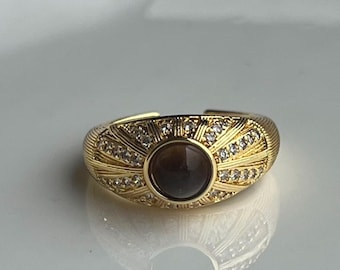 May include: Gold-toned ring with a central brown stone and surrounding clear crystals. The ring features a sunburst design with etched details. The band is adjustable, and the ring is set against a white background.