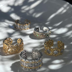 May include: An assortment of open-ended rings in gold and silver tones, adorned with intricate designs and gemstones. The rings are displayed on a white surface, with shadows adding depth to the composition. The rings feature various patterns and textures.