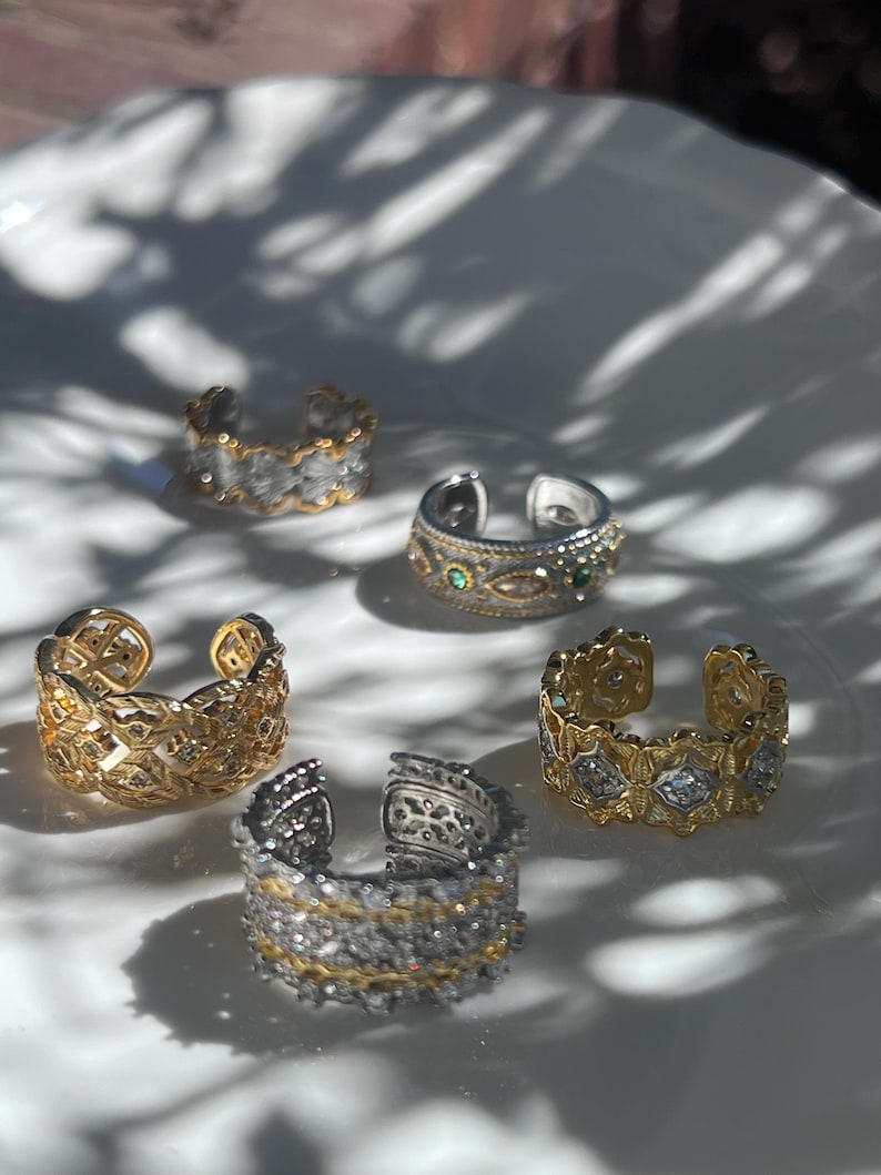 May include: An assortment of open-ended rings in gold and silver tones, adorned with intricate designs and gemstones. The rings are displayed on a white surface, with shadows adding depth to the composition. The rings feature various patterns and textures.