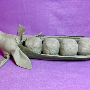 Puede incluir: Una vaina de guisante verde de peluche con cuatro guisantes redondos grises en su interior. La vaina est&aacute; hecha de tela y tiene una costura cosida en el centro. Los guisantes tambi&eacute;n est&aacute;n hechos de tela y tienen un patr&oacute;n de X cosido en ellos.