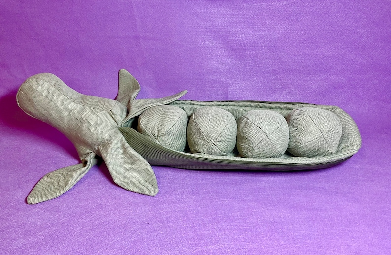 Puede incluir: Una vaina de guisante verde de peluche con cuatro guisantes redondos grises en su interior. La vaina est&aacute; hecha de tela y tiene una costura cosida en el centro. Los guisantes tambi&eacute;n est&aacute;n hechos de tela y tienen un patr&oacute;n de X cosido en ellos.