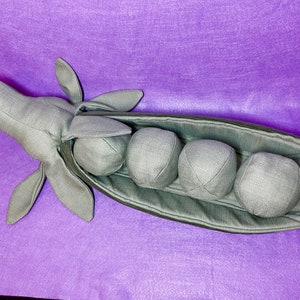 Puede incluir: Una vaina de guisante verde de peluche con cinco guisantes redondos en su interior. La vaina est&aacute; abierta y los guisantes son visibles. La vaina est&aacute; hecha de tela suave y tiene una costura cosida en el centro.